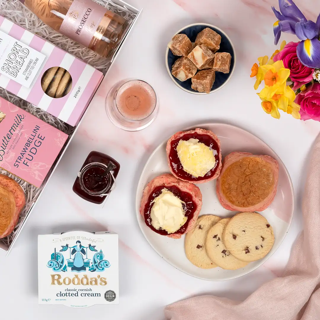 Birds eye view of Mother's Day cream tea hamper showing opened heart shaped jar of jam next to a plate of pink scones topped with jam and cream sitting next to cornish biscuits